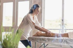 Portrait of a young masseuse performing a leg massage on a woman lying on a massage table. photo