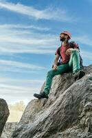 Smiling man sitting on top of rocky mountain, with climbing safety gear. photo