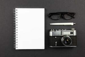 Modern office workplace flat lay with blank spiral paper notebook mockup, retro camera, pen and reading glasses on dark black background. Top view. Copy space. photo