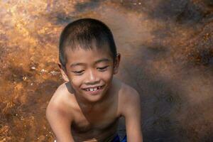 asiático joven chico linda sonriente y encantador jugando agua en río en antecedentes agua superficie y amaneceres, niño jugando agua, estilo de vida de asiático niños, concepto de vida y sostenibilidad. foto