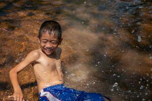 asiático joven chico linda sonriente y encantador jugando agua en río en antecedentes agua superficie y amaneceres, niño jugando agua, estilo de vida de asiático niños, concepto de vida y sostenibilidad. foto