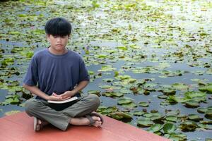 Asian boy meditating on floor at lotus pond and sky sunlight background,close up boy sitting,boy blue shirt,children relax,concept meditation and relaxation,garden thai beautiful background,riverside. photo
