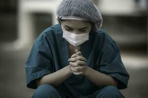 Tired depressed female asian scrub nurse wears face mask blue uniform sits on hospital floor,Young woman doctor stressed from hard work photo