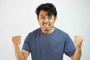 Angry aggressive young asian man wearing t shirt navy color shouting out loud with ferocious expression and mad raising fists frustrated photo