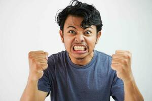 Angry aggressive young asian man wearing t shirt navy color shouting out loud with ferocious expression and mad raising fists frustrated photo