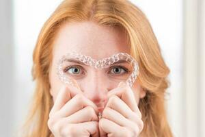 Dental care.Smiling girl with red hair holding heart shaped aligners photo