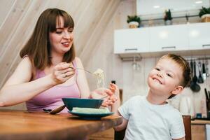 Cheerful child eating pasta in the kitchen with mom photo
