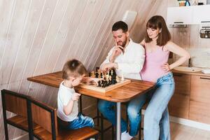 Pregnant woman watching dad and son play chess at the table photo