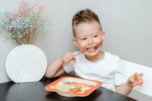 contento niño comiendo delicioso pasta a el mesa en el cocina foto