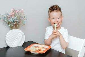 contento niño comiendo delicioso pasta a el mesa en el cocina foto