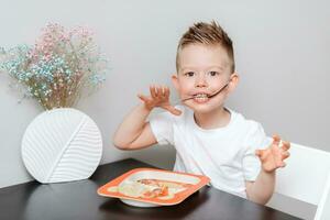 contento niño comiendo delicioso pasta a el mesa en el cocina foto