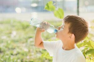 Cute boy sitting on the grass drinks water from a bottle in the summer at sunset. Child quenches thirst on a hot day photo