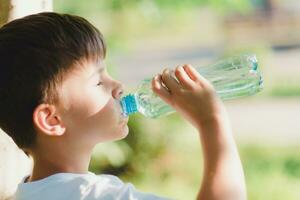 Cute boy sitting on the grass drinks water from a bottle in the summer at sunset. Child quenches thirst on a hot day photo