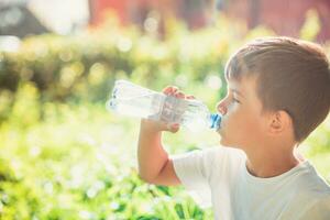 Cute boy sitting on the grass drinks water from a bottle in the summer at sunset. Child quenches thirst on a hot day photo