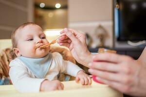 Mother feeds happy little daughter fruit puree from a spoon. First food photo