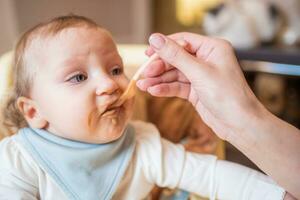 Mother feeds happy little daughter fruit puree from a spoon. First food photo