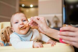 Mother feeds happy little daughter fruit puree from a spoon. First food photo