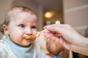 contento madre alimentación su linda pequeño hija Fruta puré desde un cuchara. primero comida foto