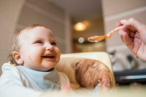 Happy mother feeding her cute little daughter fruit puree from a spoon. First food photo