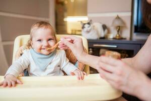 mamá alimenta su pequeño hija Fruta puré desde un cuchara. primero comida foto