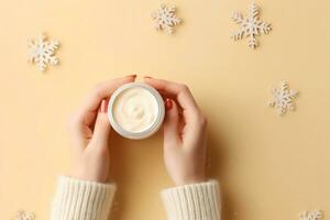 Winter season skin care cosmetics concept. First person top view photo of woman's hands in knitted sweater small cream jar and snowflakes on isolated pastel beige background. generative ai.