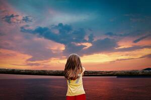 little cute girl seen from behind leaning against a wooden fence is looking at stunning sunset on the sea photo