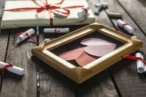 Flat lay romantic background. Giftbox, rolled wish papers, and wooden frame with paper hearts on dark wood board. photo