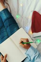 High angle view to young woman lying in bed and writing with pen on notebook. Wintertime. Christmas holidays. A woman making plans for the new year photo