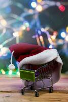 Christmas shopping X-mas sale concept. Shoping cart and Santa cap against decorated with festive lights Christmas tree. Christmas spendings photo