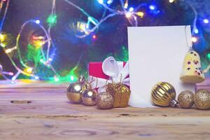 Christmas tree decorations and gift box with a blank card on a wooden table. Xmas toys with blank paper over Christmas tree and lights bokeh. Holiday decoration. Xmas background photo