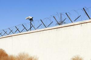 Security camera behind barbed wire fence stretched around prison walls. Security, surveillance camera watching area photo