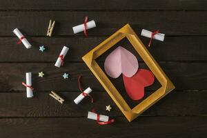 Flat lay romantic background. Giftbox, rolled wish papers, and wooden frame with paper hearts on dark wood board. photo