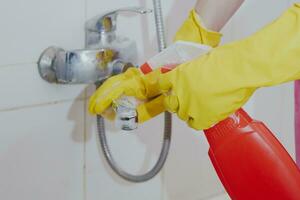 Housewife cleaning bathroom tap and shower Tap. Maid in yellow protective gloves washing dirty bath tap. Hands of woman washing bath photo