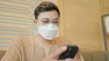 A happy man wearing a face mask sits typing a message on his smartphone. video