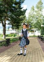 alegre gracioso niña con un sin dientes sonrisa en un colegio uniforme con blanco arcos en colegio patio trasero. espalda a escuela, septiembre 1. contento alumno con un mochila. primario educación, elemental clase. foto