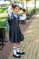 alegre gracioso niña con un sin dientes sonrisa en un colegio uniforme con blanco arcos en colegio patio trasero. espalda a escuela, septiembre 1. contento alumno con un mochila. primario educación, elemental clase. foto