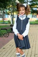 alegre gracioso niña con un sin dientes sonrisa en un colegio uniforme con blanco arcos en colegio patio trasero. espalda a escuela, septiembre 1. un contento alumno. primario educación, elemental clase. retrato de un estudiante foto