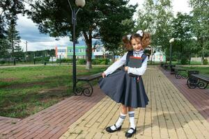 Girl with backpack, school uniform with white bows and stack of books near school. Back to school, happy pupil, heavy textbooks. Education, primary school classes, September 1 photo