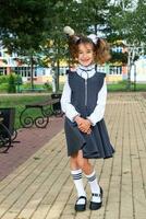 Cheerful funny girl with a toothless smile in a school uniform with white bows in school yard. Back to school, September 1. A happy pupil. Primary education, elementary class. Portrait of a student photo