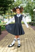 Cheerful funny girl with a toothless smile in a school uniform with white bows in school yard. Back to school, September 1. A happy pupil. Primary education, elementary class. Portrait of a student photo