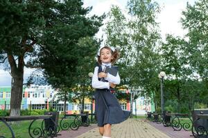 alegre gracioso niña con sin dientes sonrisa en colegio uniforme con blanco arcos corriendo en colegio patio trasero. espalda a escuela, septiembre 1. contento alumno con mochila. primario educación, elemental clase. movimiento foto