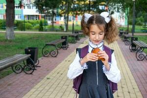 Girl with a backpack eating pie near school. A quick snack with a bun, unhealthy food, lunch from school. Back to school. Education, primary school classes, September 1 photo