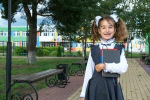 Girl with backpack, school uniform with white bows and stack of books near school. Back to school, happy pupil, heavy textbooks. Education, primary school classes, September 1 photo