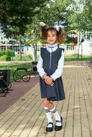 Cheerful funny girl with a toothless smile in a school uniform with white bows in school yard. Back to school, September 1. A happy pupil. Primary education, elementary class. Portrait of a student photo