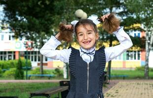 Cheerful funny girl with a toothless smile in a school uniform with white bows in school yard. Back to school, September 1. A happy pupil. Primary education, elementary class. Portrait of a student photo