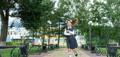 alegre gracioso niña con sin dientes sonrisa en colegio uniforme con blanco arcos corriendo en colegio patio trasero. espalda a escuela, septiembre 1. contento alumno con mochila. primario educación, elemental clase. movimiento foto