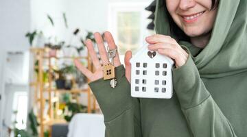 A happy woman in her house holds in her hands a miniature figure of a house and key in the interior. Dream house project, real estate purchase, insurance, mortgage, rent, reservation photo