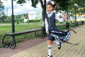 alegre gracioso niña con sin dientes sonrisa en colegio uniforme con blanco arcos corriendo en colegio patio trasero. espalda a escuela, septiembre 1. contento alumno con mochila. primario educación, elemental clase. movimiento foto