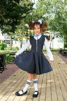 Cheerful funny girl with a toothless smile in a school uniform with white bows in school yard. Back to school, September 1. A happy pupil. Primary education, elementary class. Portrait of a student photo