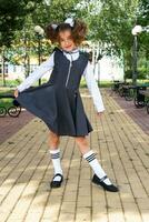 alegre gracioso niña con un sin dientes sonrisa en un colegio uniforme con blanco arcos en colegio patio trasero. espalda a escuela, septiembre 1. un contento alumno. primario educación, elemental clase. retrato de un estudiante foto